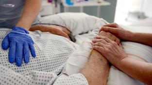 Eine Krankenschwester kümmert sich um den Patienten. Eine Angehörige hält ihm die Hand. (Foto: picture alliance/Patrick Seeger/dpa)