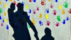 Ein Mann mit einem Kind auf dem Arm und einem an der Hand wirft einen Schatten auf eine mit bunten Handabdrücken bemalte Wand (Foto: picture alliance/Peter Kneffel/dpa)