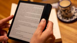 Ein Mann hält einen E-Book-Reader in der Hand, während im Hintergrund zahlreiche Papierbücher auf dem Tisch liegen. (Foto: picture alliance/dpa | Monika Skolimowska)