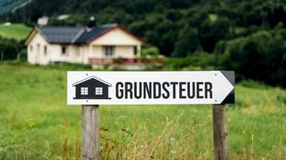Wegweiser aus Holz in einer Wiese vor einem Haus mit der Aufschrift: Grundsteuer (Foto: picture alliance / CHROMORANGE | Michael Bihlmayer)