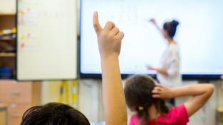 Ein Schüler meldet sich per Handzeichen während eine Lehrerin auf eine digitale Schultafel im Klassenraum einer 4. Klasse einer Grundschule schreibt (Foto: picture alliance/dpa | Julian Stratenschulte)