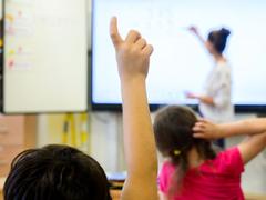 Ein Schüler meldet sich per Handzeichen während eine Lehrerin auf eine digitale Schultafel im Klassenraum einer 4. Klasse einer Grundschule schreibt (Foto: picture alliance/dpa | Julian Stratenschulte)