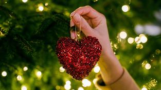 Eine Frau hängt eine herzförmige Christbaumkugel in einen Baum mit Lichterkette (Foto: picture alliance / Westend61 | Yuliia Blazhuk)