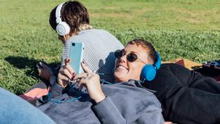 Zwei Frauen liegen auf einer Wiese und hören über Kopfhörer etwas auf ihrem Smartphone. (Foto: IMAGO / Westend 61)