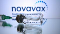 Impfdosen mit Impfstoff zur Injektion mit einer Kanüle. Im Hintergrund das Logo "Novavax". (Foto: IMAGO / Sven Simon)