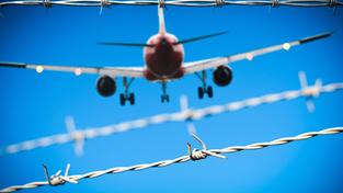 Symbolbild Abschiebung von Straftätern. Ein Flugzeug fliegt am Himmel hinter einem Stacheldrahtzaun. (Foto: IMAGO / Bihlmayerfotografie)