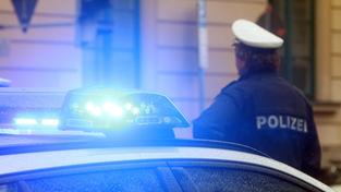 Ein Polizist in Uniform steht vor einem Einsatzwagen der Polizei mit Blaulicht (Foto: IMAGO / Ralph Peters)